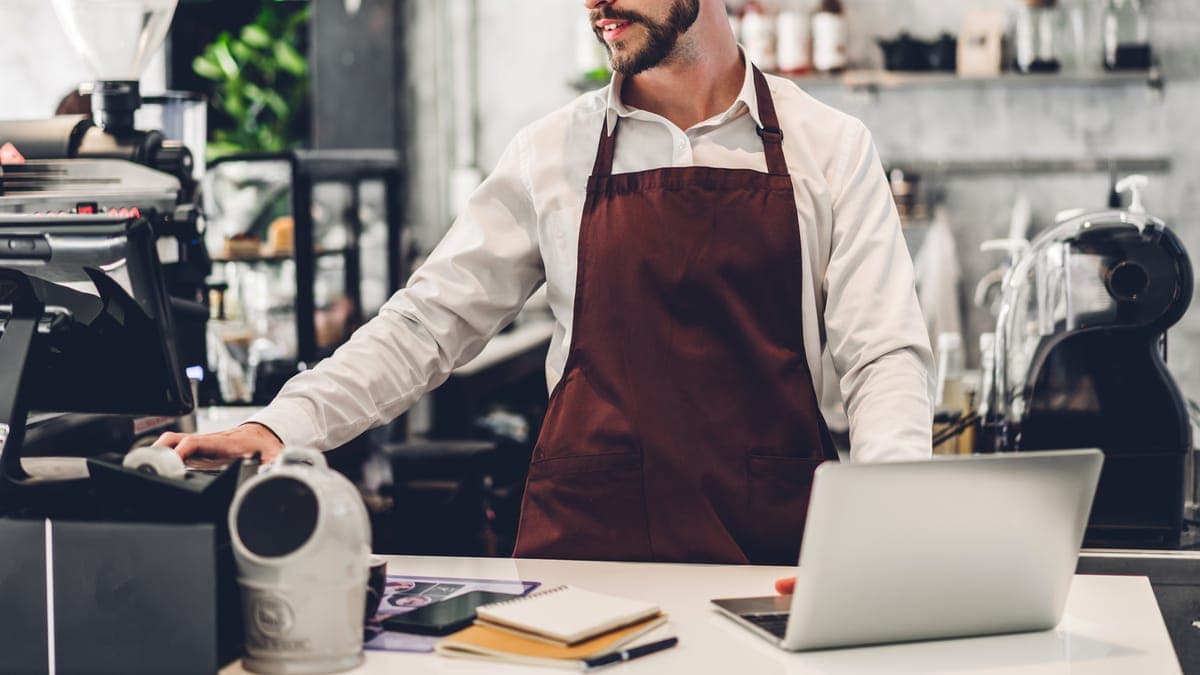 Barista in a brown apron at the cafe counter with a laptop and notebooks