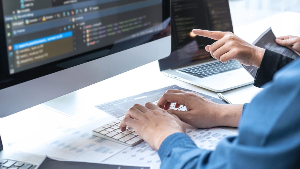 Developers reviewing code on a large monitor and laptop at an office desk